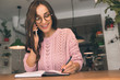 © LIGHTFIELD STUDIOS - happy woman in eyeglasses woman talking on smartphone while writing in textbook at table in cafe