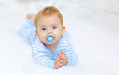 charming blue-eyed baby 7 month old lies in bed in a striped bodysuit and sucks a pacifier