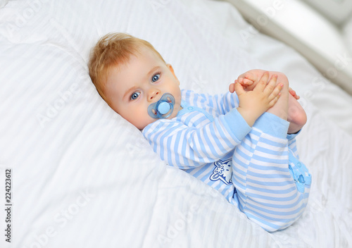 Fototapeta Charming blue-eyed baby 7 month old lies in bed in a striped bodysuit and sucks a pacifier