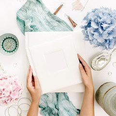 Wall Mural - Woman hands hold family wedding photo album, pastel colorful hydrangea flower bouquet, turquoise blanket, decoration, fashion accessories on white background. Flat lay, top view.