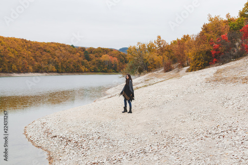 Fotografía Young brunette woman wearing grey poncho walking alone near the lake in autumn