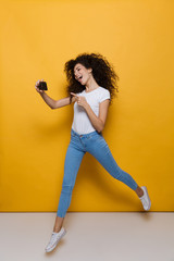 Sticker - Full length photo of european woman 20s with curly hair holding smartphone and taking selfie photo, isolated over yellow background