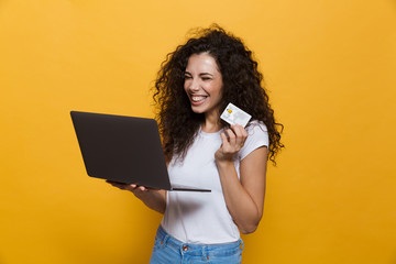 Sticker - Photo of gorgeous woman 20s wearing casual clothes holding black laptop and credit card, isolated over yellow background