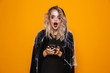 Blond woman wearing black costume and halloween makeup holding mobile phone, isolated over yellow background
