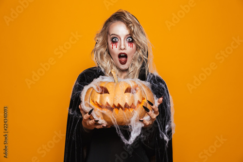 Fotomural Magician woman wearing black costume and halloween makeup holding carved pumpkin