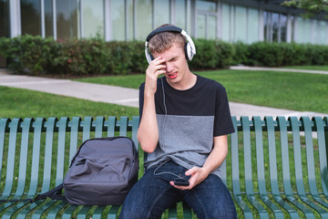 Wall Mural - Depressed teenager sitting on bench outside of his school while listening to music with his headphones.