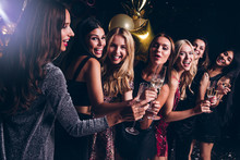 Pure Celebration. Beautiful Young Women In Evening Gown Holding Champagne Glasses And Looking At Camera With Smile While Celebrating In Nightclub