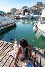 Happy Tourist Girl On Holiday Trip To Heraklion, Crete, Greece. Young Woman Traveller Enjoyes Sunshine Sitting On Wooden Walkway Of Heraklion Venetian Port Marina