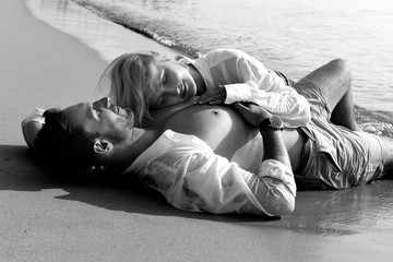 Beautiful happy couple in love lying on a beach on sand in a white shirts are in love smiling. Handsome bearded men and sexy blonde women in water in wet clothes having fun kissing, and laughing