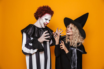 Image of witch woman and joker man wearing black costume and halloween makeup playing around, isolated over yellow background