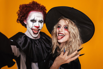 Poster - Photo of joyful witch woman and clown man wearing black costume and halloween makeup taking selfie, isolated over yellow background