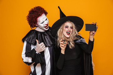 Poster - Photo of horrific witch woman and joker man wearing black costume and halloween makeup taking selfie on smartphone, isolated over yellow background