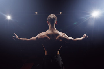 Rear view of healthy muscular man with arms outstretched black background light searchlights