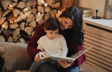 Pretty mother and her little daughter reading book in the evening at home with cozy and christmas decoration. Beautiful woman and cute little girl have literature. Family, hygge and people concept