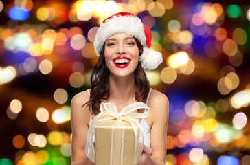 Wall Mural - christmas, holidays and people concept - happy smiling young woman with red lipstick in santa hat holding present over festive lights background