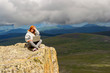 © Aleksandr Kondratov - A pretty red-haired woman in sports clothes will work as a freelancer while traveling on the edge of a high mountain on a dangerous cliff at the bottom of which the lake and fields with plants