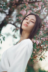 Wall Mural - Gorgeous asian woman with perfect skin creartive art make-up wearing trendy white japanese kimono standing agaist flower background in the garden. Close-up fashion portrait outdoors. Spring blossom of