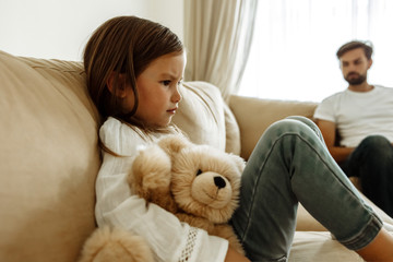 Childhood. Education. Problem. Sad little girl is hugging her teddy bear, her father in the background