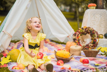 Little girl lying and playing in a tent, children's house wigwam in park Autumn portrait of cute curly girl. Harvest or Thanksgiving. autumn decor, party, picnic. Child in yellow dress with apple 