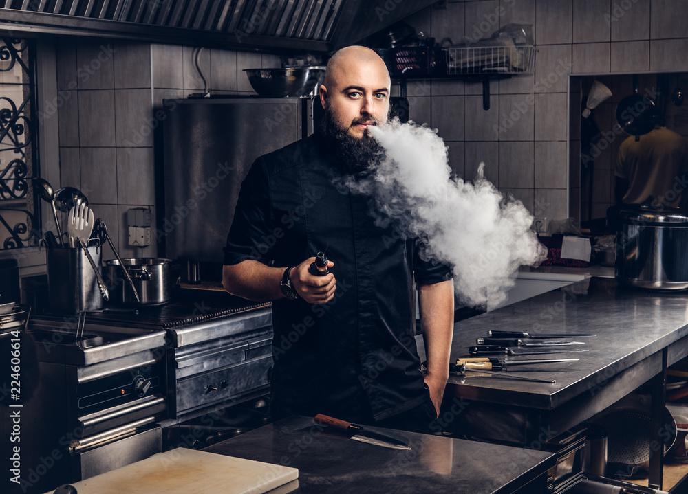 Bearded chef in black uniform smoking e-cigarette while standing in the  kitchen. Stock Photo