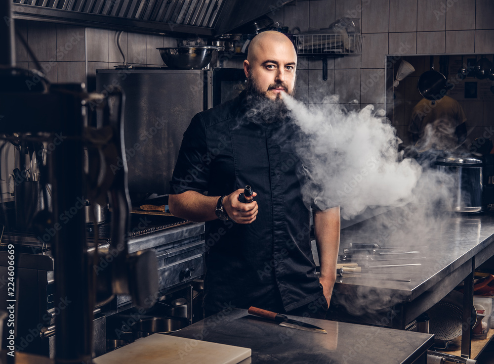 chef cigarette Bearded chef in black uniform smoking e-cigarette while standing in the  kitchen. Stock Photo