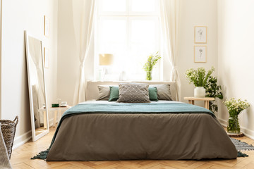 Beige, green and gray bedroom interior in a tenement house with a bed against a sunny window and bunches of wild flowers. Real photo.