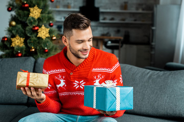 smiling young man sitting on sofa and holding gift boxes near christmas tree at home