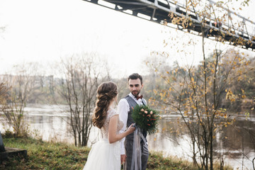 Happy young smiling bride and groom stand near the suspension bridge and the river. Wedding photos in an interesting place