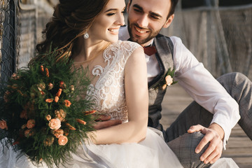 Happy young smiling bride and groom are sitting on the suspension bridge. Sunny wedding photos in an interesting place