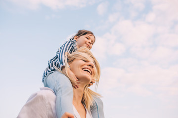 Bottom view of cute little daughter on a piggy back ride with her smiling mother on sky background. Loving woman and her little girl playing in the park. Mom and kid have fun outside. Motherhood