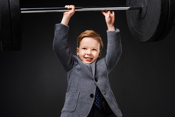 strong smiling little boy in suit lifting barbell isolated on grey