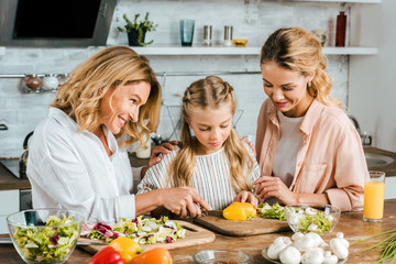 Wall Mural - concentrated little child cutting vegetables for salad with mother and grandmother at home