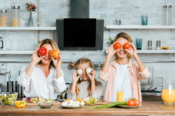 Wall Mural - playful little child covering eyes with food with mother and grandmother and looking at camera while cooking