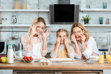 Wall Mural - child with mother and grandmother looking at camera through dough cutters while cooking