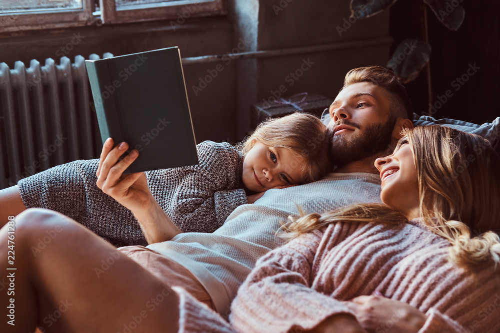 Mom, dad and daughter reading storybook together while lying on bed. - obrazy, fototapety, plakaty 