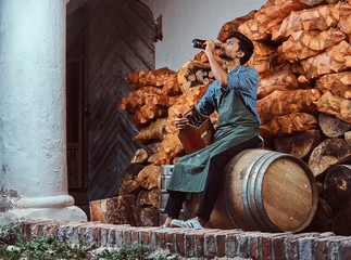 Wall Mural - Tired brewer in apron sitting on a barrel and drinks craft beer relaxing after work.