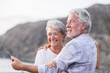 © simona - concept of vacation, technology, tourism, travel and people - happy senior couple with cell phone phone on pebble beach laughing and joking hugging and making photo. White hair