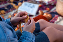 A Young Woman In A Jeans Jacket And  Knitting  Yellow Hat With Needle And Natural Wool, Sitting On A Plaid With A Picnic Basket, Apples.Concept Of A Freelancer Work In The Open Air