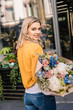 © LIGHTFIELD STUDIOS - smiling attractive girl standing with wrapped bouquet near flower shop and looking at camera