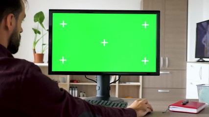 Side view of man sitting in front of computer with big green screen mock-up chroma typing on the keyboard. He is in the living room of his house