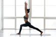 © fizkes - Young sporty woman practicing yoga doing Warrior one exercise, Virabhadrasana 1 pose, working out wearing sportswear pants, bra, at indoor at yoga studio, full length side view, white window