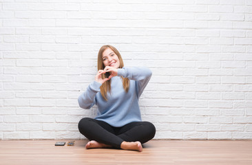 Sticker - Young adult woman sitting on the floor in autumn over white brick wall smiling in love showing heart symbol and shape with hands. Romantic concept.