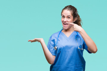 Sticker - Young brunette doctor girl wearing nurse or surgeon uniform over isolated background amazed and smiling to the camera while presenting with hand and pointing with finger.