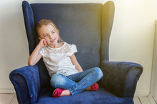 Sweet Little Girl In Jeans And White T-shirt At Home Sitting On Modern Cozy Blue Chair Relaxing In White Living Room. Childhood, Schoolchildren, Youth, Relax Concept