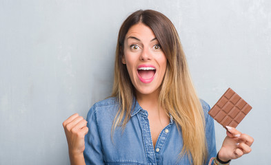 Young adult woman over grey grunge wall eating chocolate bar screaming proud and celebrating victory and success very excited, cheering emotion