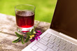 Glass Of Berry Juice With Meadow Flower Near Laptop On An Wooden Table On Bright Sunny Day Or At Dawn. Grape or Cherry Juice In Misted And Covered With Drops Of Glass. Concept Of Vacation Office Work.