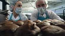 Medium Shot Of Two Confectionery Workers, Man And Woman, Pushing Baking Rack With Fresh Cupcakes And Talking To Each Other When Working At Factory