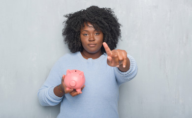 Wall Mural - Young african american woman over grey grunge wall holding piggy bank pointing with finger to the camera and to you, hand sign, positive and confident gesture from the front