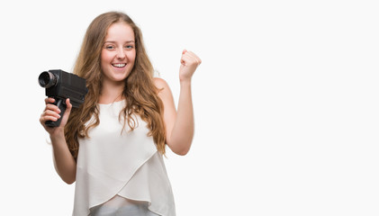 Wall Mural - Young blonde woman holding super 8 video camera screaming proud and celebrating victory and success very excited, cheering emotion