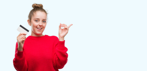 Wall Mural - Young blonde woman holding credit card very happy pointing with hand and finger to the side